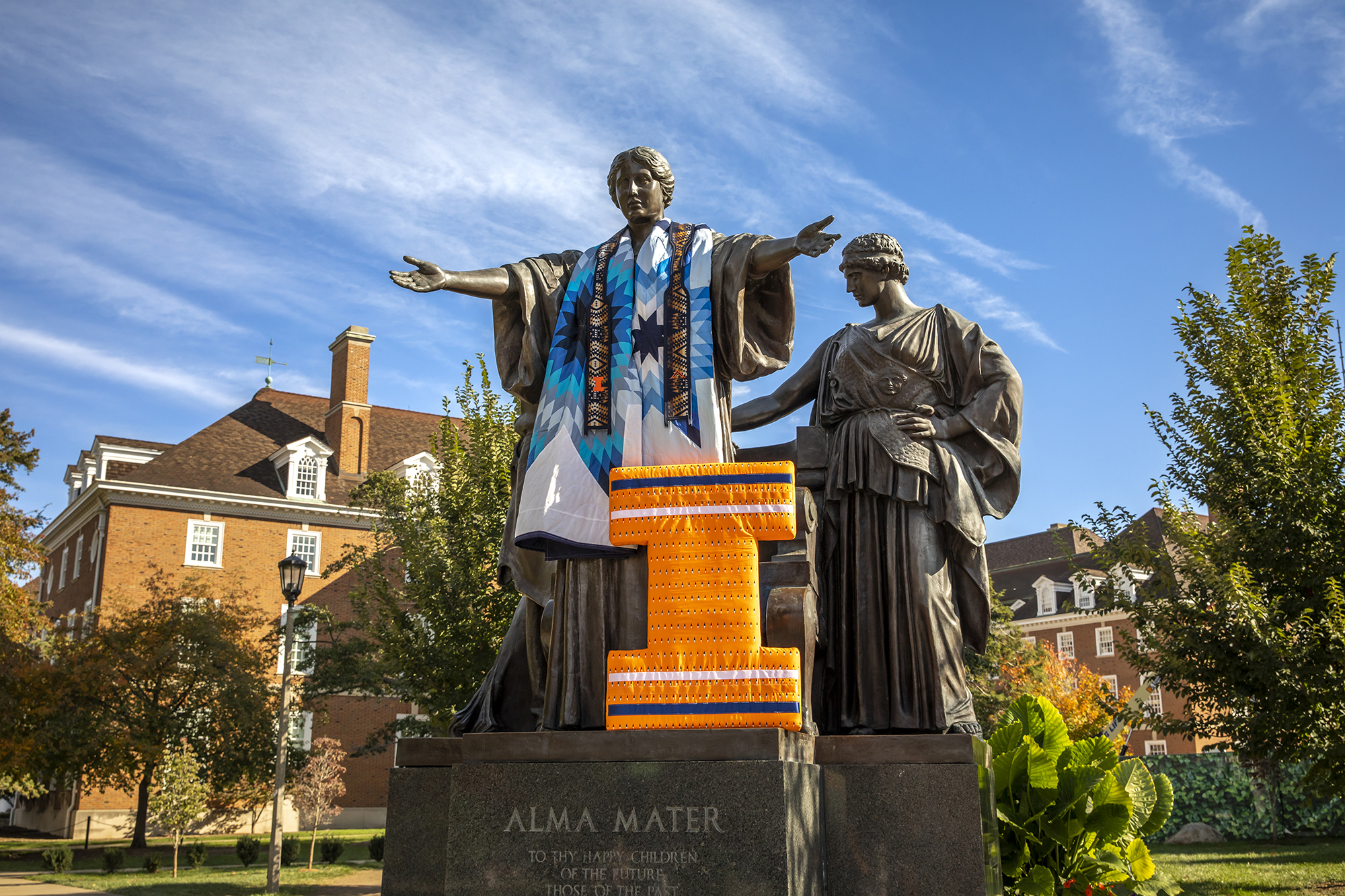 Picture of Alma Mater decorated in a Native blanket with different shades of blue and white, as well as a graduation stole with Native/geometric designs in darker colors. There is also a big &quot;I&quot; in orange, blue and white leaning against Alma Mater. There are clear blue skies and the Illini Union in the background, alongside trees and plants.
